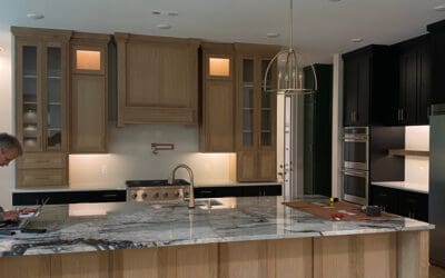 Far view of redone modern kitchen. Island with wooden cabinets, marble countertops and floor lights. There is a man working on the counter